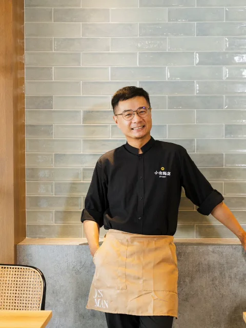 小金穿著麵店制服、繫著圍裙，靠在店裡的牆邊。